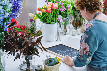 Side view of unrecognizable female florist writing on black chalkboard preparing bright flower shop for Christmas holidays