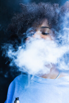 Modern relaxed African American female with curly hair exhaling fume through nose while smoking on street