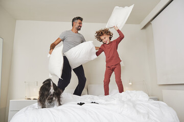 Playful father and son fighting on bed with soft pillows while jumping and having fun