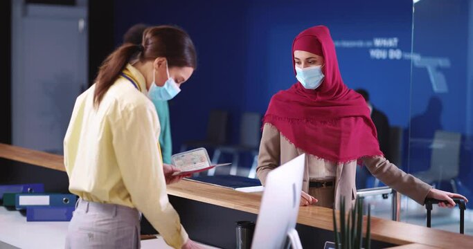 Female Security Agent In Face Mask Servicing Diverse Multi-cultural Customers On Check-in Desk Reading Travel Ticket Before Flight. Airport Terminal. Registration.