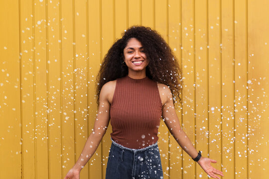 Joyful African American female with curly hair scattering white confetti in city while having fun and looking at camera