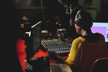 Back view of ethnic female in headphones sitting at table with mixing console and speaking in microphone while working in broadcast studio