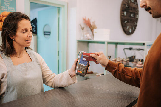 Positive female seller standing at counter and holding terminal while customer paying with credit card using NFC technology in confectionery cafe
