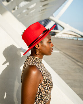 Side view of dreamy African American female in red trendy hat standing with closed eyes in city on sunny day