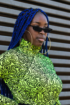 Side view of unemotional black female with blue braided hair and in bright clothing standing on street and looking away