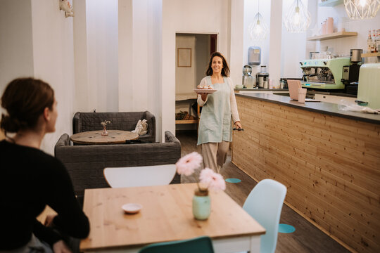 Positive female confectionery owner carrying plate with sweets while serving dessert for guest in cozy cafe