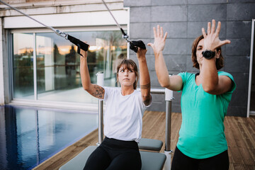 Female in Navasana doing yoga with support of personal instructor while balancing on pilates reformer with straps on terrace