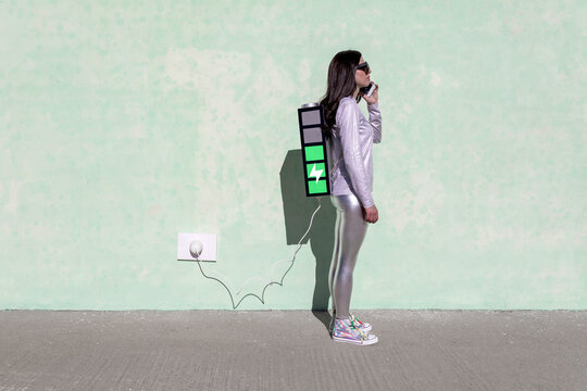 Side view of young fit female in trendy outfit standing on street with creative backpack connected to charger and having phone conversation