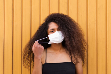 Positive African American female with curly hair wearing white protective mask standing near urban building in city during coronavirus pandemic and looking at camera