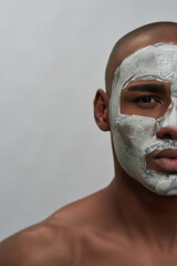 Half face portrait of shirtless young african american man looking at camera using facial clay mask, posing isolated over gray background