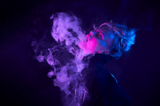 Side view of young cool female smoking vape in neon light in dark studio with eyes closed