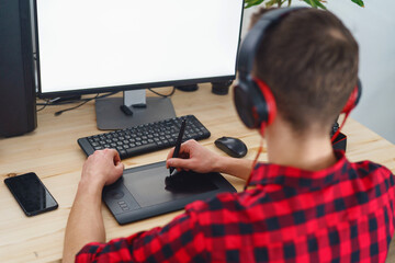 Rear view, adult young guy wearing headphones, using pen tablet, doing retouching in front computer at home. photographer processes photos. blank white computer screen. Freelancer. Remote work. Blogs