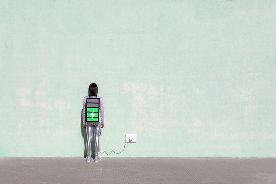 Back view of unrecognizable female with long dark hair in stylish outfit standing on street near wall with battery shaped backpack connected to charger