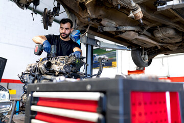 Low angle of focused male mechanic with electric screwdriver fixing engine of car while working in service
