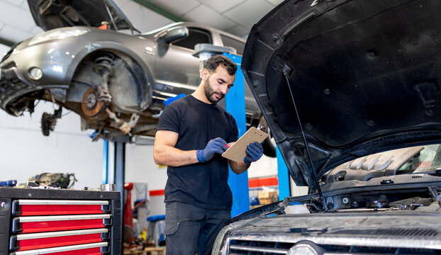 male technician analyzing chart on clipboard while working in modern automobile workshop and examining car