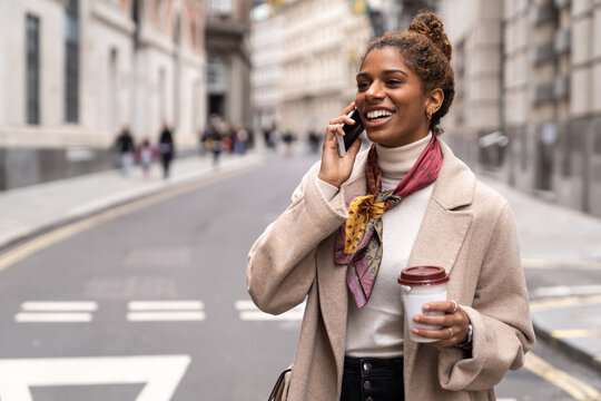 Side view of young ethnic lady in stylish coat walking on city square and having phone conversation in London