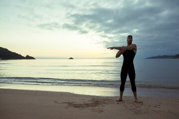 Back view of fit male swimmer in swimsuit and cap standing on seashore and stretching arms before training
