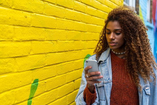 Content young African American female millennial with long curly hair in casual outfit browsing on smartphone while walking on cobblestone street near colorful building with abstract graffiti