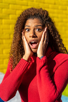 Astonished African American female teenager with long curly hair in red sweater standing near colorful graffiti wall with opened mouth and hands on cheeks and looking at camera