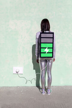 Back view of unrecognizable female with long dark hair in stylish outfit standing on street near wall with battery shaped backpack connected to charger