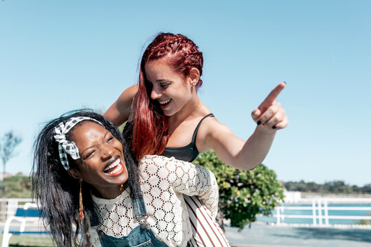 Laughing African American woman giving piggyback ride to girlfriend pointing away while having fun together in sunny summer day on street