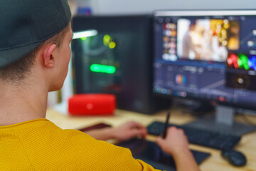young guy videographer uses pen-tablet and special program to process video on front computer at home. Video editing. Blogs. Freelancer. Remote work. Retouching training. View from over the shoulder