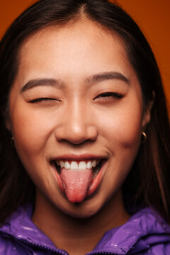 Close-up portrait of happy Asian young woman. She wears a purple jacket and is looking at camera smiling and winking against a yellow background
