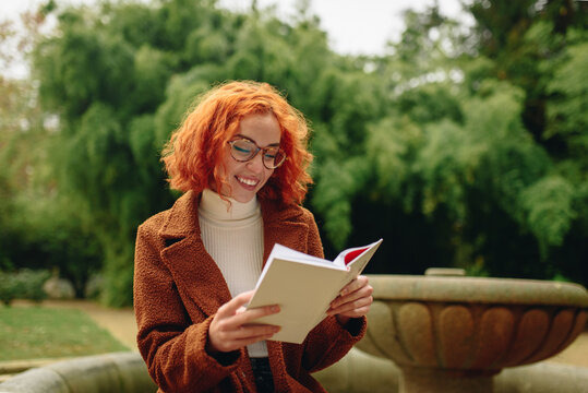Focused female with red hair sitting in park and reading interesting story in book while enjoying weekend alone