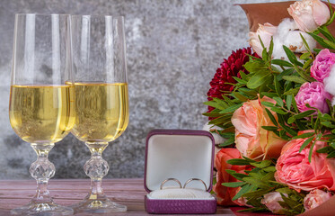 glass with champagne, bunch of flowers and wedding rings on grey background