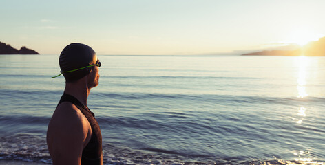 Side view of confident male swimmer in cap and goggles standing on beach on background of sea under sunset sky and looking away