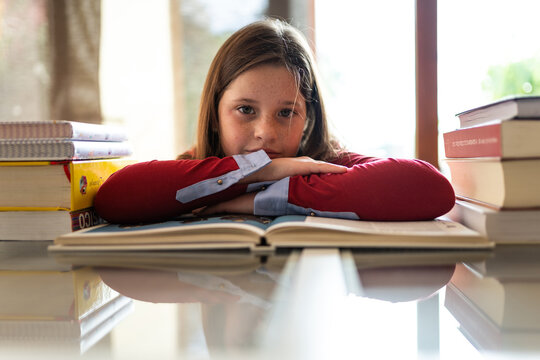 Cute diligent girl sitting at table leaning on arms near pile of textbooks and looking at camera while doing homework assignment