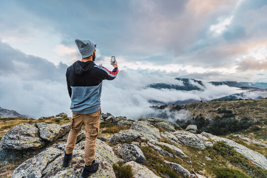 Back view of male hiker standing on rock and taking picture of spectacular highland landscape on smartphone while enjoying adventure