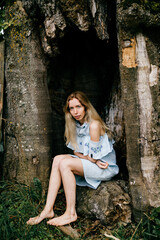 Young attractive barefoot blonde girl in blue romantic dress sitting in the old tree