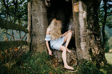 Young attractive barefoot blonde girl in blue romantic dress sitting in the old tree