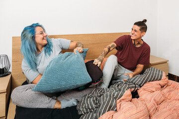 Cheerful plus size woman with dyed hair laughing and hitting tattooed girlfriend with pillow while sitting on bed and having fun together at home