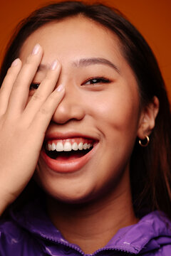 Close-up portrait of cheerful Asian young woman. She wears a purple jacket and is looking at camera smiling with hand on face against a yellow background