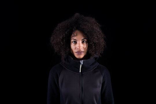 Young African American female with curly hair wearing black clothes looking at camera while standing against black background