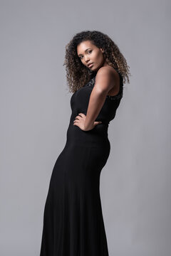 Seductive young plus size African American female model with long curly hair wearing elegant black dress standing on studio looking at camera against gray background