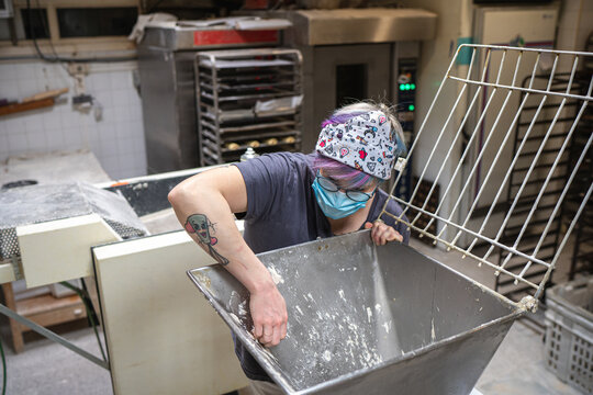 Female baker in protective mask cleaning metal baking machine from dough while working in bakehouse