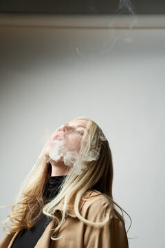 Tranquil female in stylish outfit standing in studio and smoking vape with eyes closed