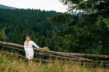 Young attractive blonde girl in white dress with embroidery posing with spikelets bouquet over picturesque landscape