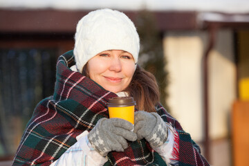 European woman in woollen gloves and checkered scarf holds a yellow paper cup of coffee on the winter city street. Hot and warming beverage in winter season