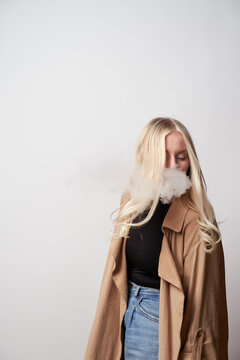 Tranquil female in stylish outfit standing in studio and smoking vape while looking down