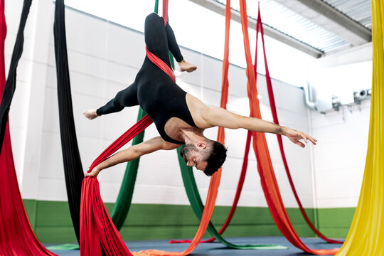 Bearded adult dancer in black leotard hanging upside down on aerial silk during rehearsal in studio