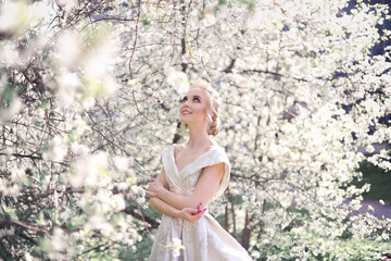 A beautiful blonde girl in a luxurious white brocade haute couture dress with a long train and open shoulders walking in a blooming orchard