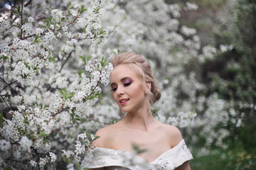 A beautiful blonde girl in a luxurious white brocade haute couture dress with a long train and open shoulders walking in a blooming orchard