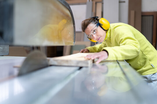 Side view of focused adult female artisan in goggles and headphones sawing wooden board with sharp circular saw in workshop