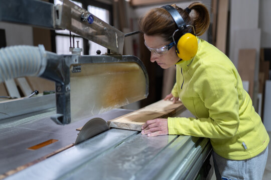 Side view of focused adult female artisan in goggles and headphones sawing wooden board with sharp circular saw in workshop