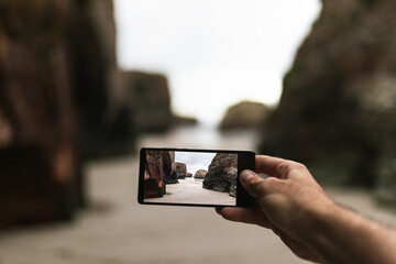 Crop anonymous male traveler taking photo of rocky sea coast on mobile phone while exploring wild beach during summer holidays
