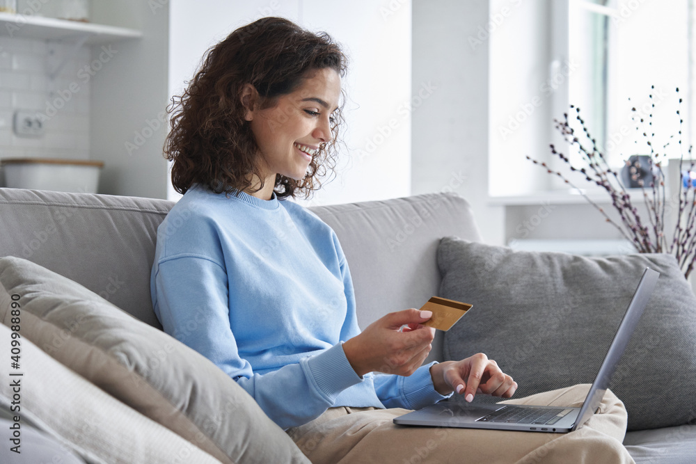 Wall mural happy hispanic young woman consumer holding credit card and laptop buying online at home. female sho